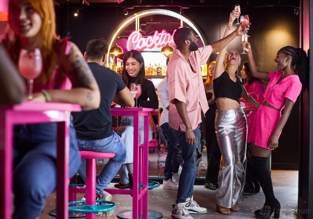 A lively bar with neon cocktail sign, pink bar stools, and guests enjoying drinks at Boom Battle Bar Birmingham Broad Street.