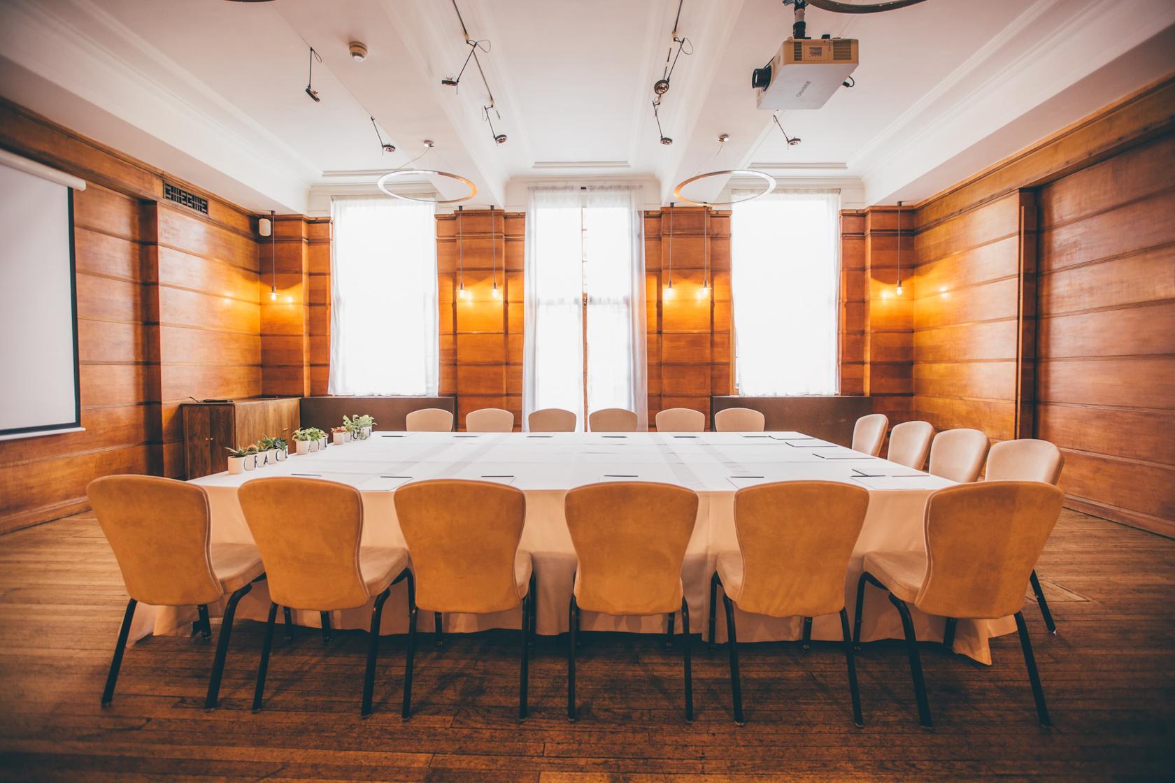 A function room with wood-paneled walls, floor-to-ceiling windows, and a U-shaped table with beige chairs at Town Hall Hotel.