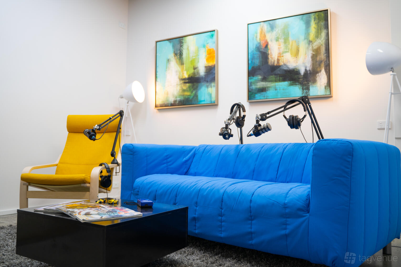 A podcast recording room with a blue sofa, microphones on adjustable arms, an armchair, and abstract art at Scroll Stop Studios.