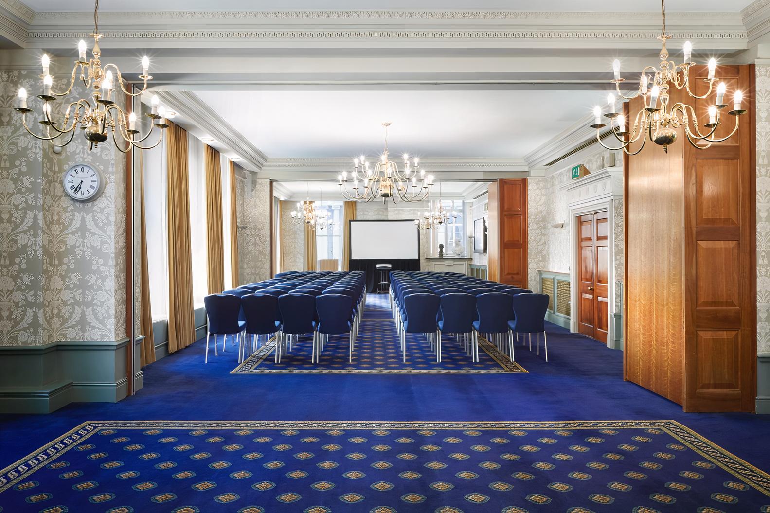 A function room with blue carpet, gold chandeliers, and rows of chairs facing a projector screen at 113 Chancery Lane.