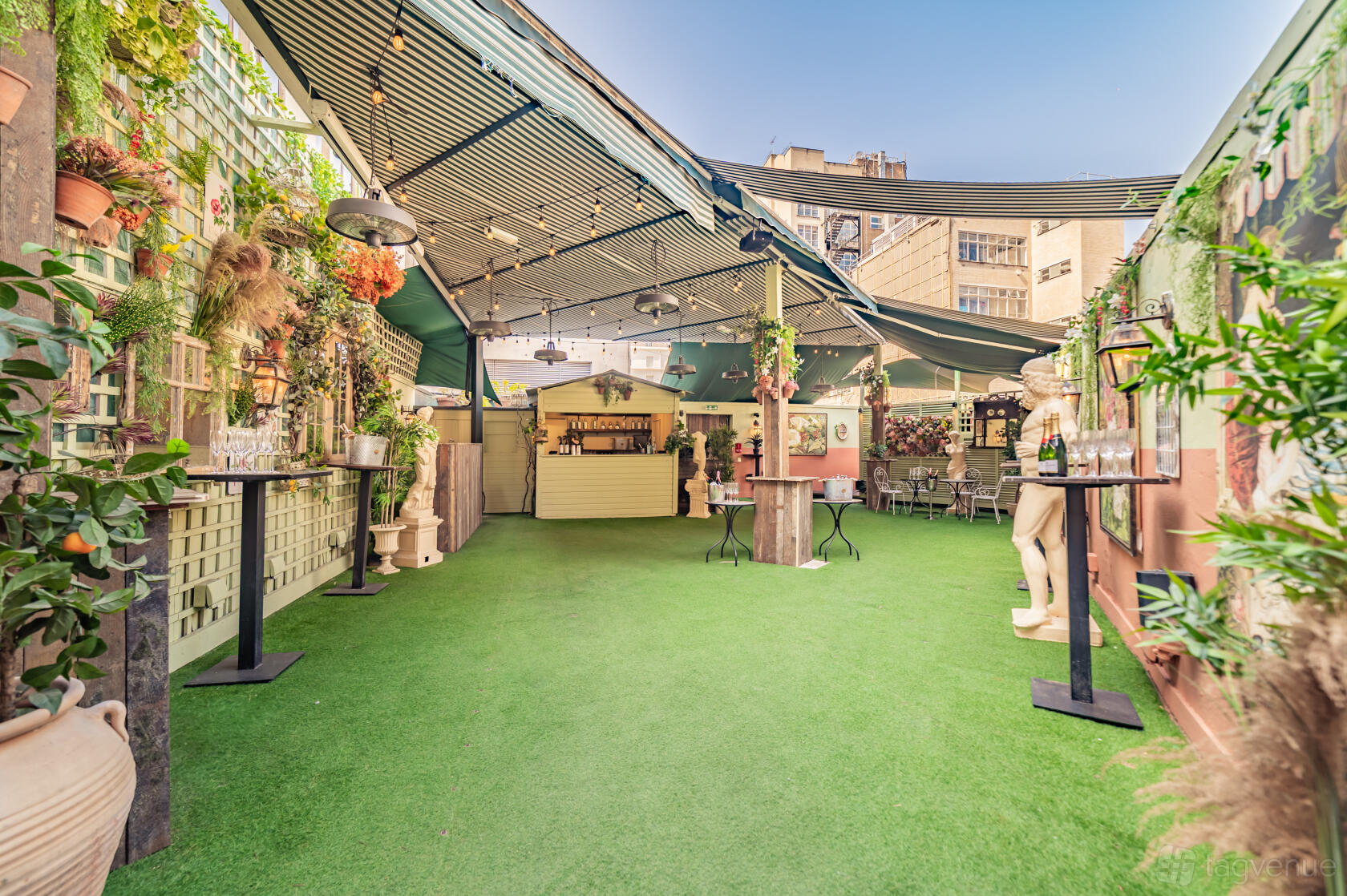 An outdoor bar with artificial grass flooring, potted plants, statues, and string lights at Secret Garden Mayfair.