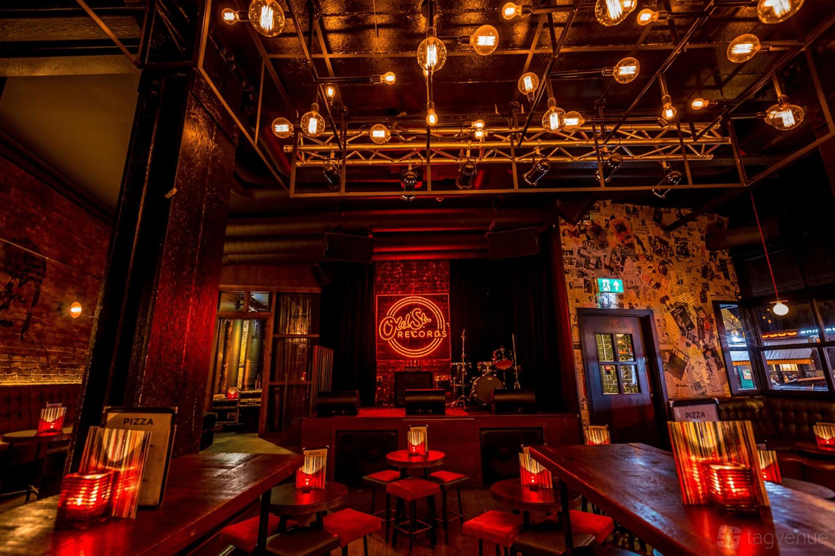 A function room with exposed brick walls, hanging Edison bulbs, and a stage with neon signage at Old Street Records.