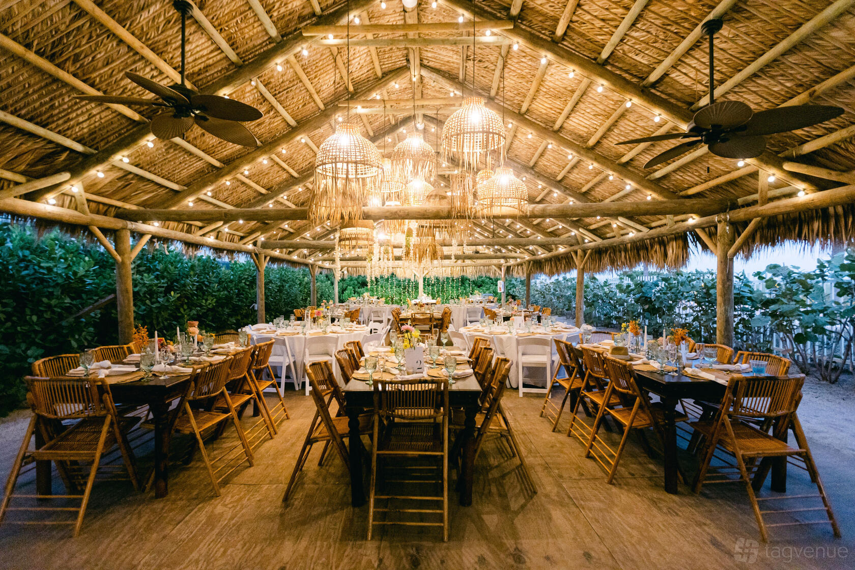 An outdoor event space with a thatched roof, woven pendant lights, and wooden tables at Palm House.