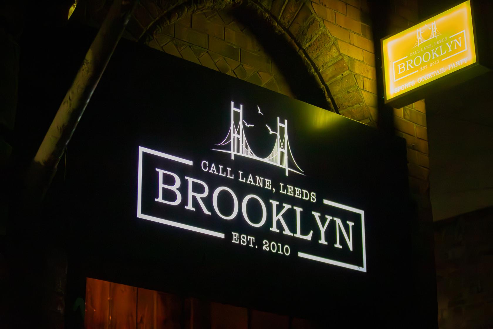 A pub with an illuminated black sign displaying a bridge logo and the name Brooklyn Bar at the entrance