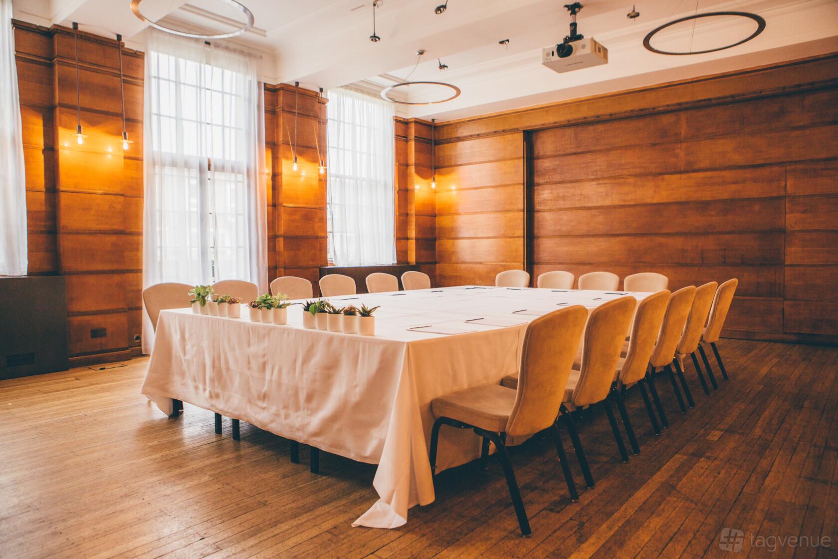A function room with large windows, wood-paneled walls, and a U-shaped table with cream chairs at Town Hall Hotel.