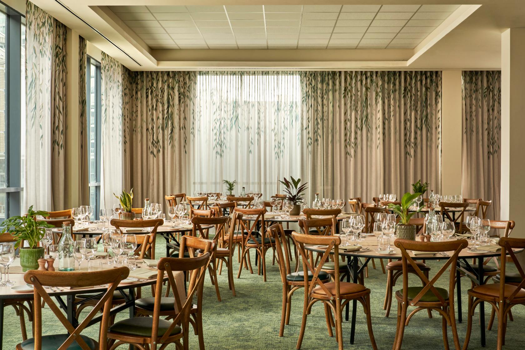 An event space in a restaurant with floor-to-ceiling windows, wooden chairs, and greenery at Treehouse Hotel Manchester.