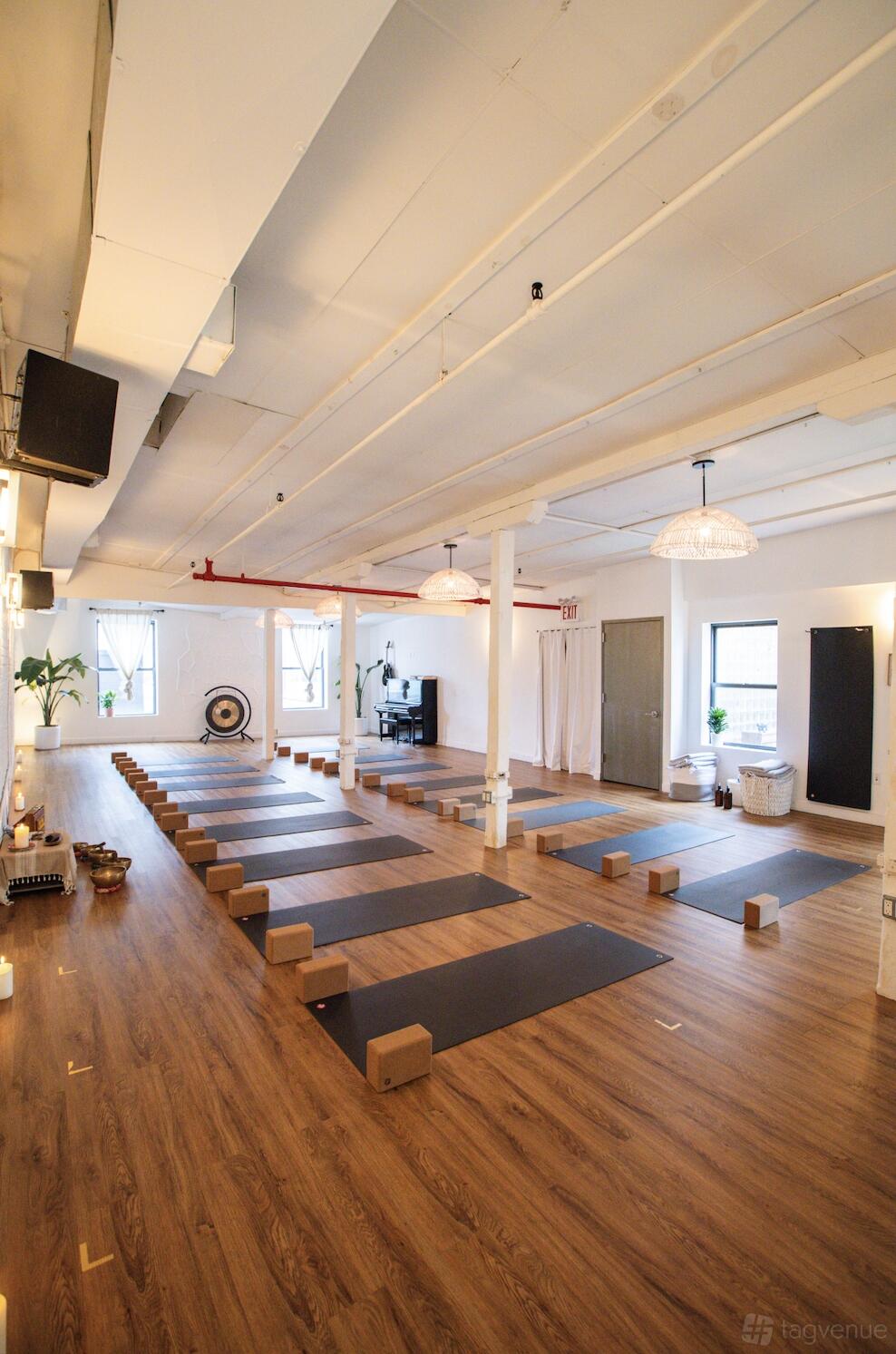 A yoga studio with wooden floors, yoga mats and blocks arranged in rows, large windows, and pendant lighting at Form + Flow Waterfront Yoga Studio.