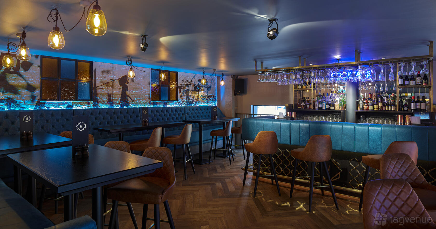 A cocktail bar with blue upholstered seating, herringbone wood floors, and a backlit bar at Manahatta Greek Street.