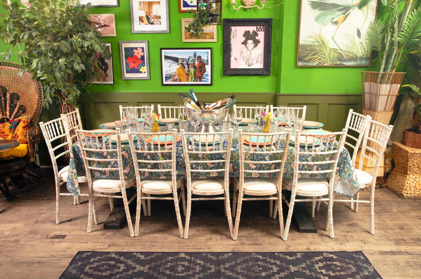 An event space in a cocktail bar with a long floral tablecloth, white chairs, and eclectic framed art at The Little Blue Door.