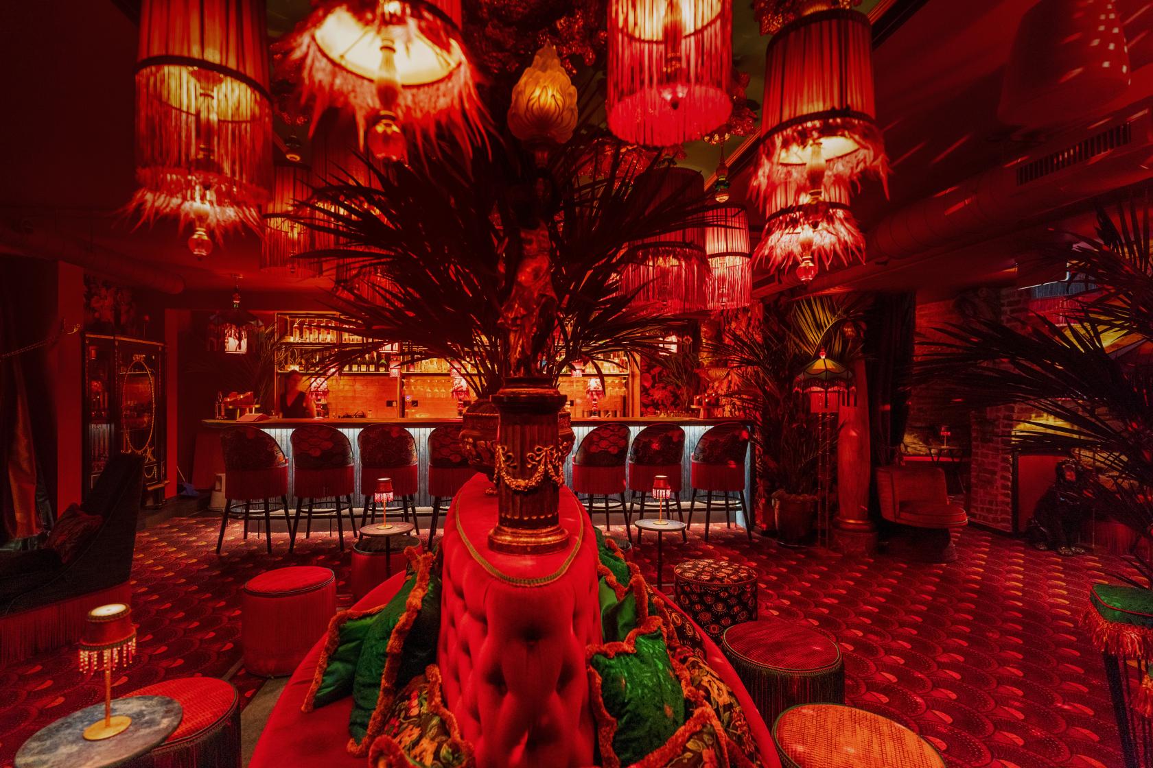 A cocktail bar with red velvet seating, fringed pendant lamps, and a long backlit bar at Zum Barbarossa.