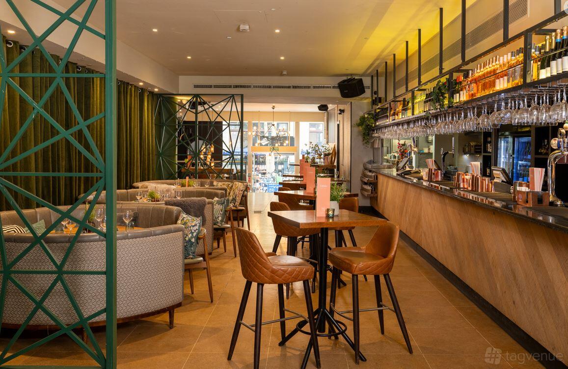 A cocktail bar with high-top tables, tan leather chairs, and an illuminated back bar at The Otherist.