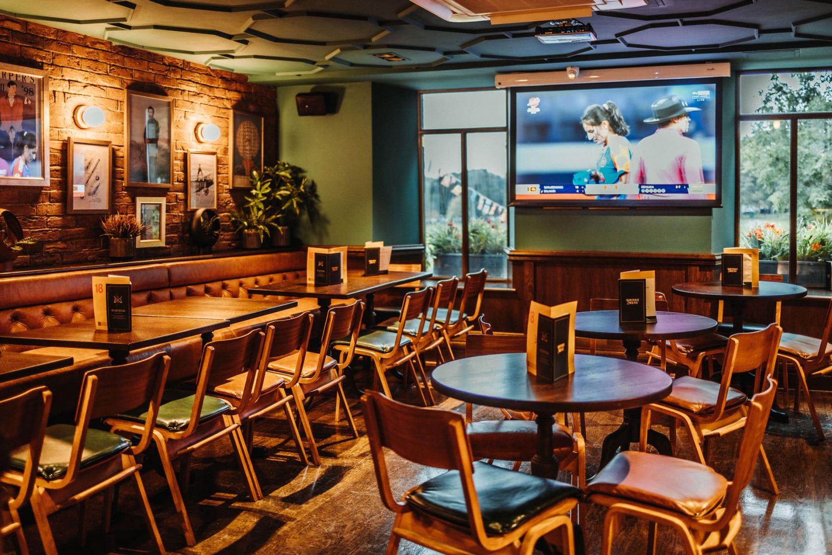 A function room in a pub with round wooden tables, cinema-style chairs, exposed brick walls, and a large TV screen at The Golf Tavern.