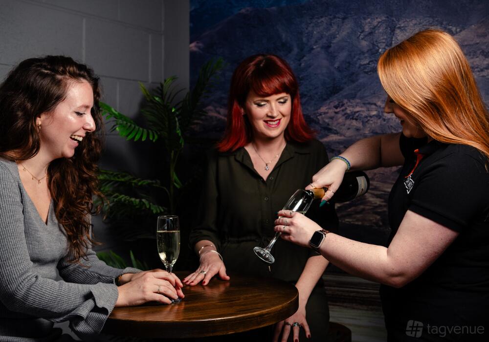 An entertainment centre with a small round wooden table, leafy plant, and people enjoying drinks at Escape Hunt Bristol.