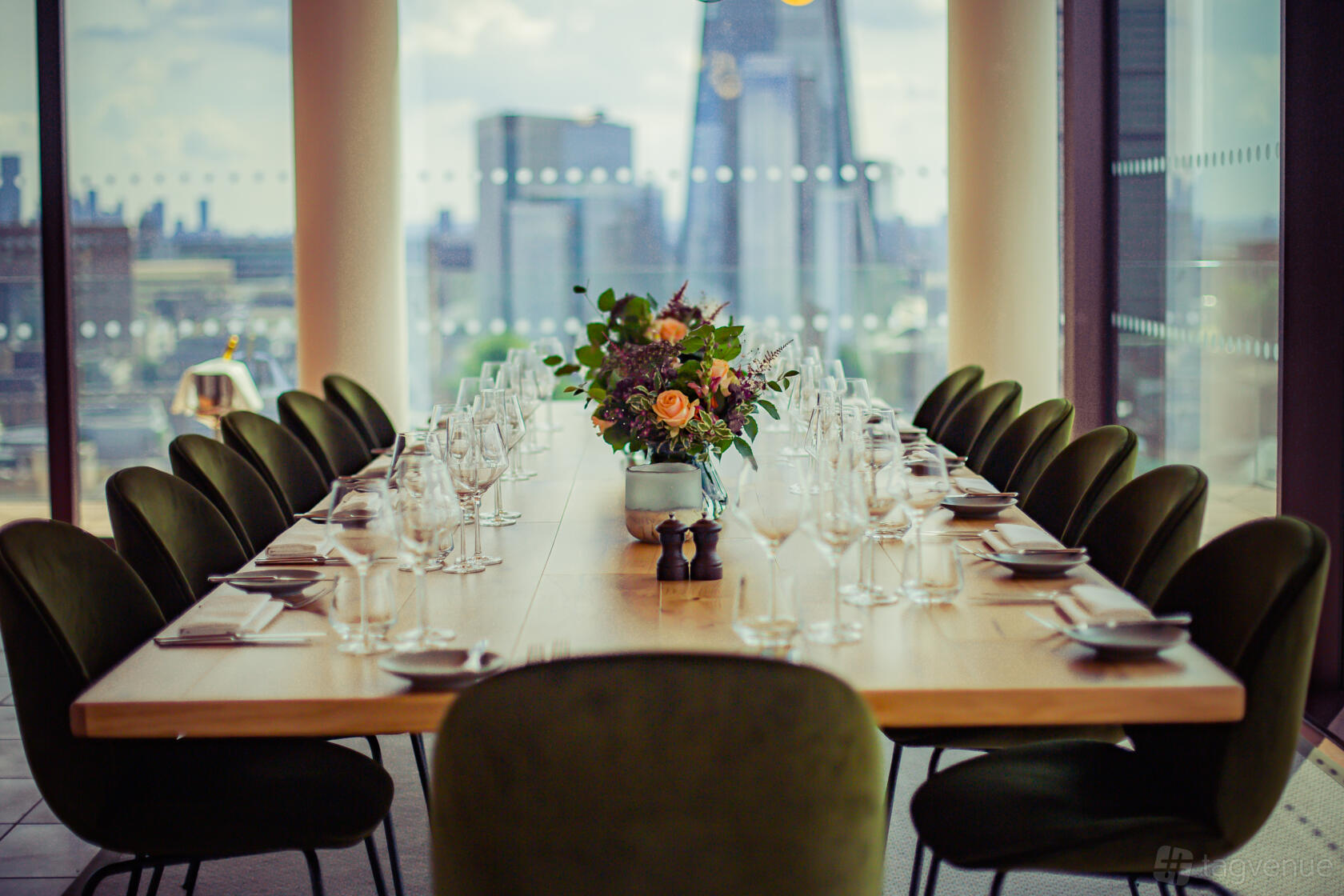 A dining room with a long wooden table, green velvet chairs, and city skyline views at Rose Court Events.
