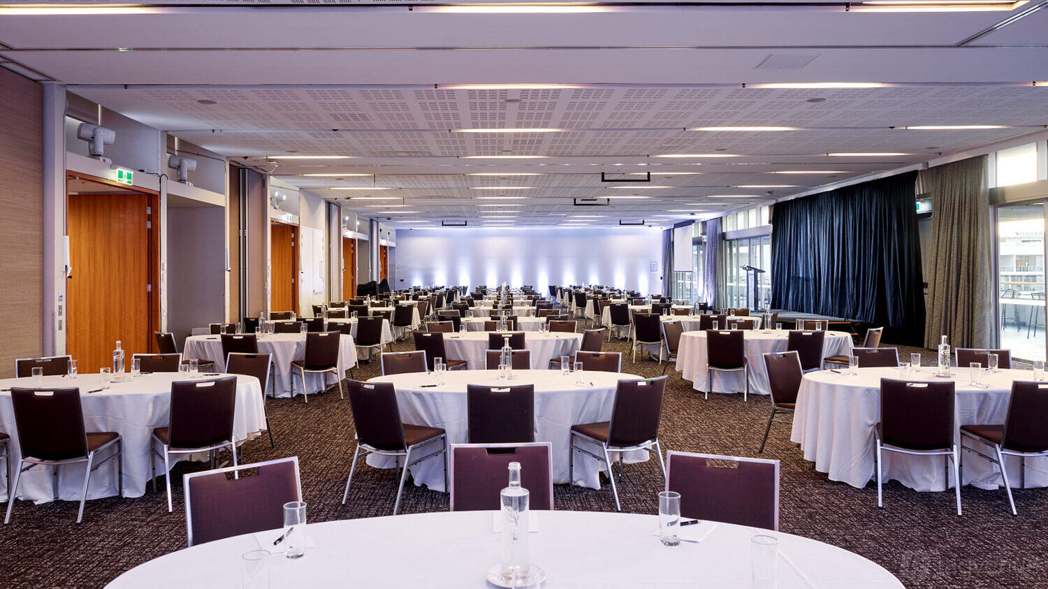 A ballroom with rows of round tables and chairs, white tablecloths, and bottled water at Aerial Function Centre.