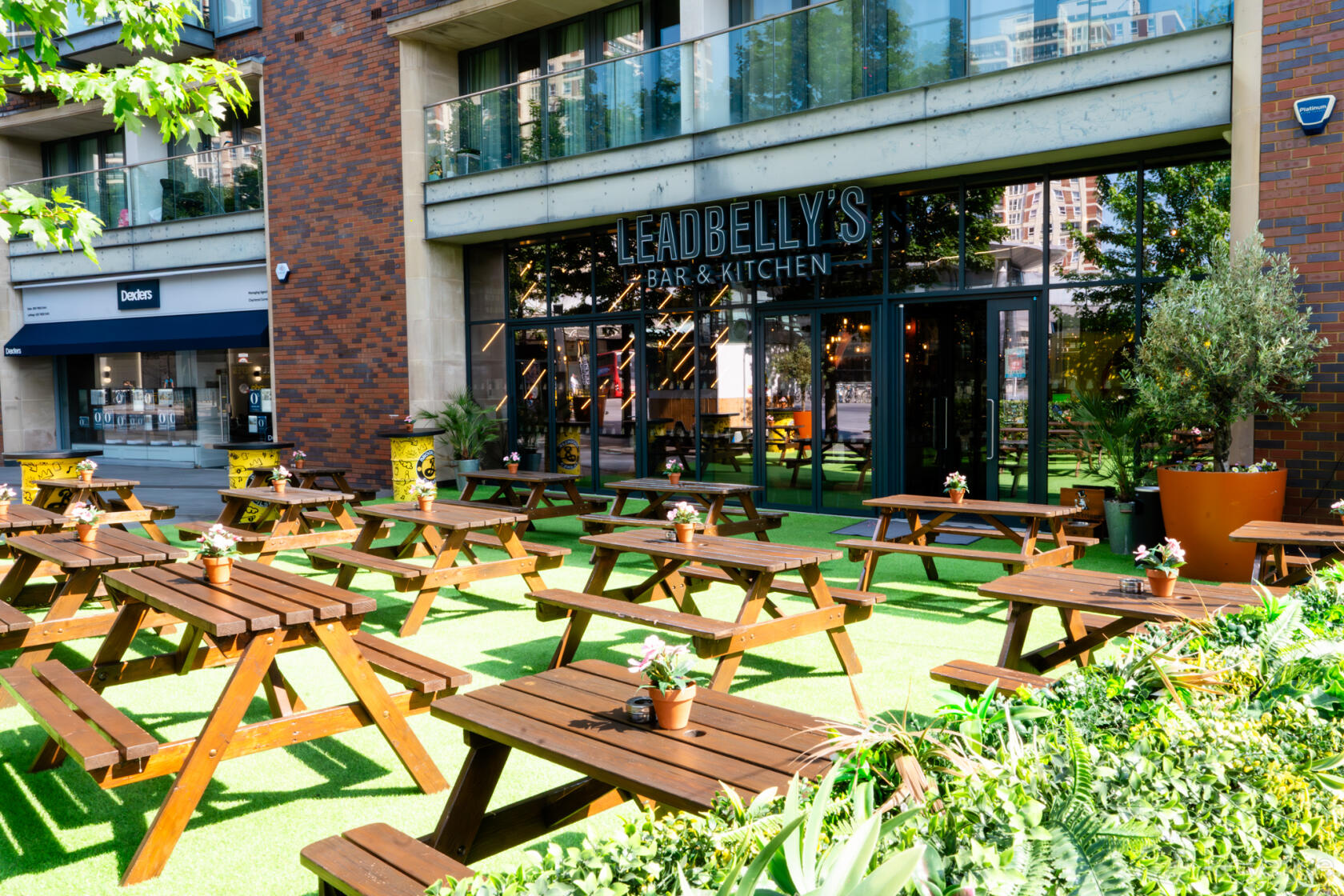 A terrace at a bar with wooden picnic tables, potted plants, and artificial turf at Leadbelly's Bar & Kitchen.