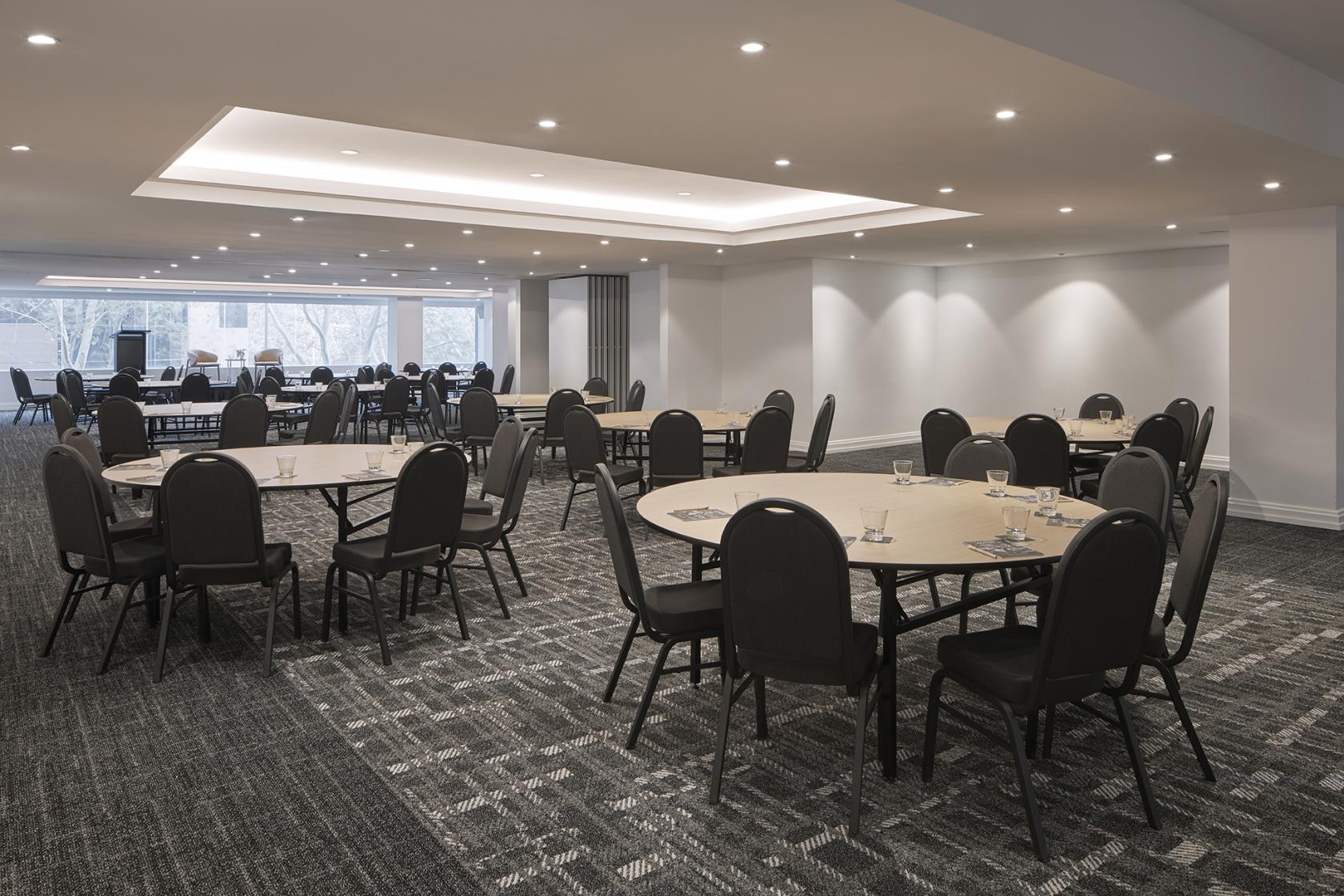 An auditorium with round tables, black chairs, carpeted flooring, and recessed ceiling lights at Rydges Sydney Central.