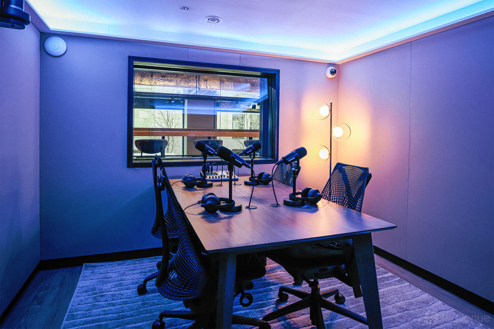 A podcast studio with four microphones, mesh chairs, soft lighting, and a window at Qube East - Cocktail Bar & Recording Studios.