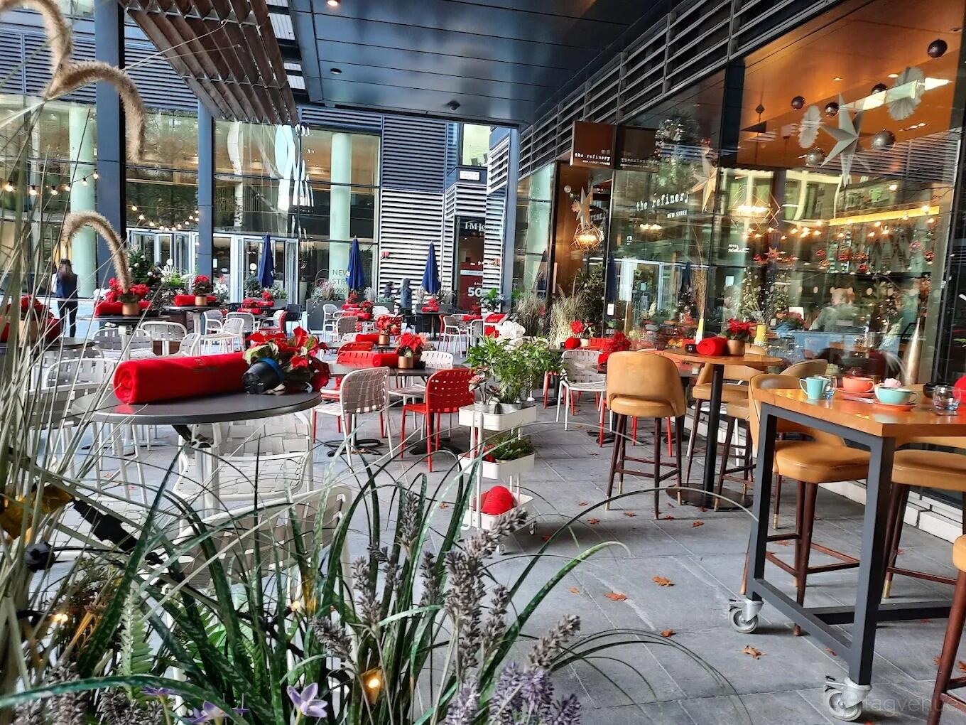 An outdoor restaurant terrace with red-accented chairs, potted plants, and glass walls at The Refinery New Street Square.