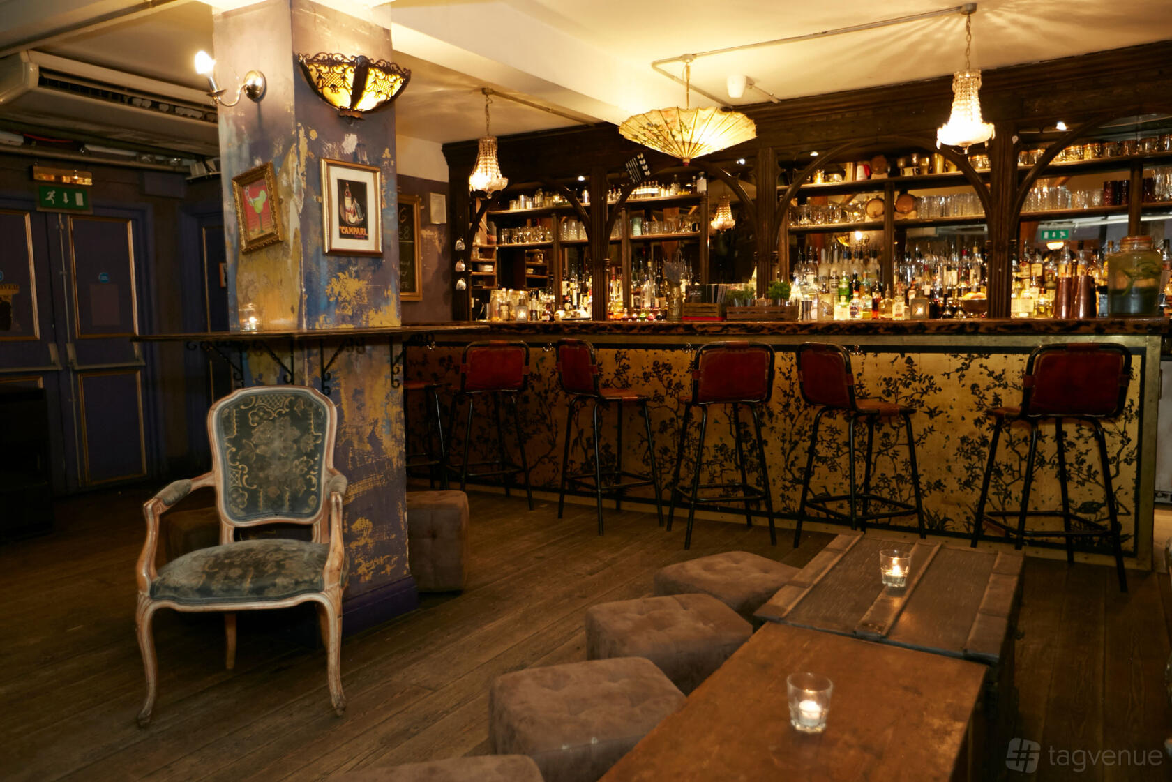 A cocktail bar with a gilded bar front, backlit shelves of bottles, red velvet bar stools, and vintage armchairs at The Lucky Pig Cocktail Bar.