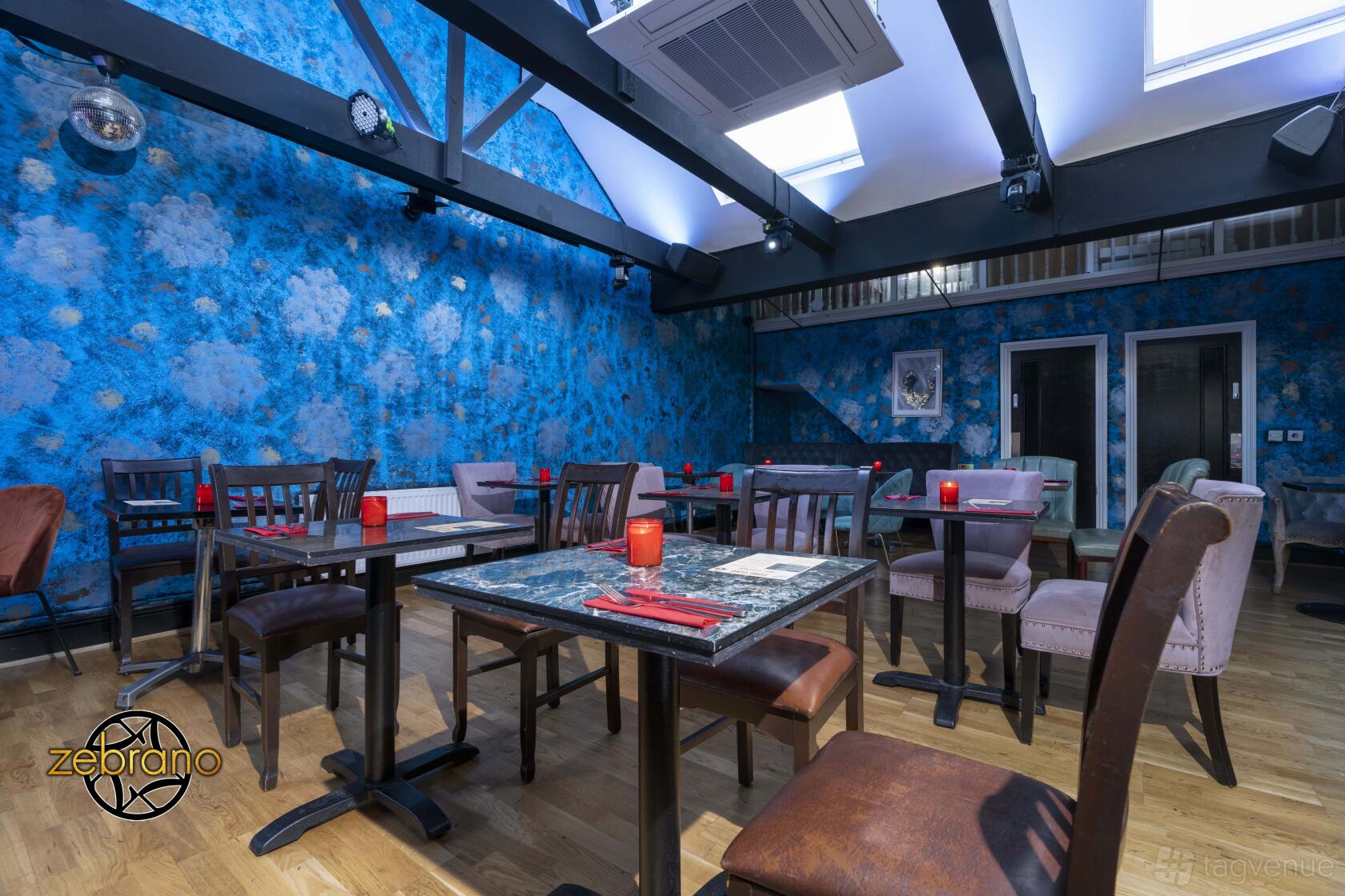 A lounge in a bar with blue patterned walls, exposed beams, and mixed wooden tables and chairs at Soho Zebrano.
