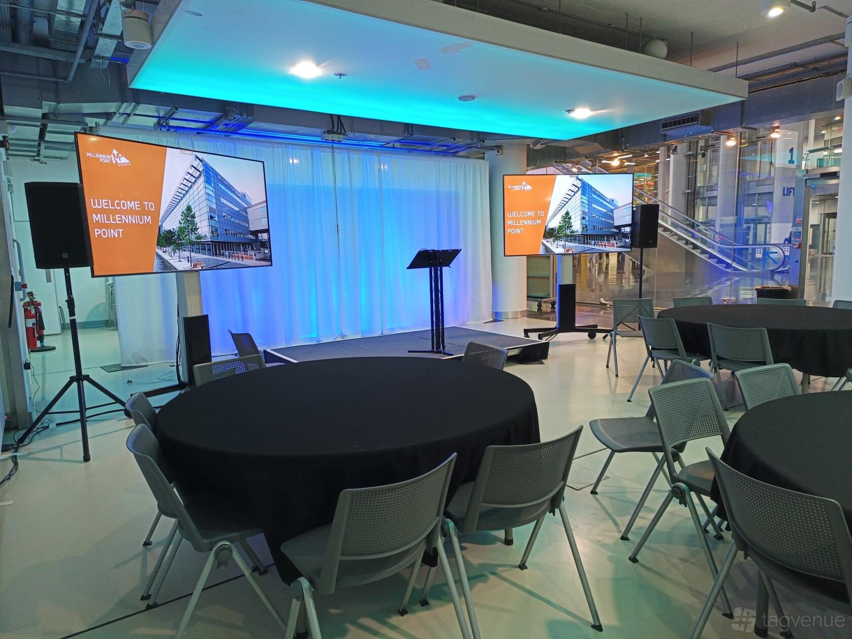 A meeting room with round tables, grey chairs, two large display screens, and a lectern at Millennium Point.