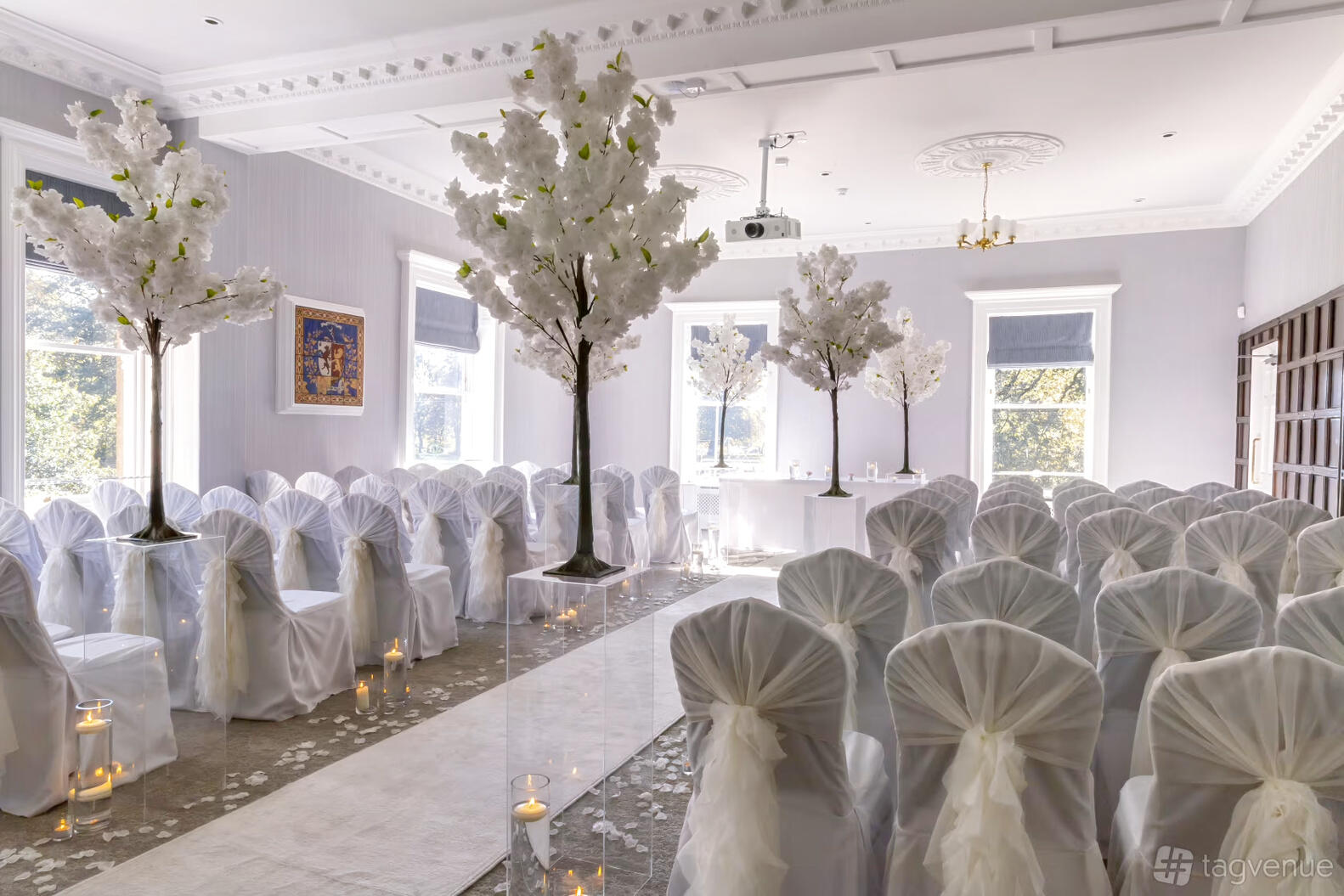 An event space with white chair covers, aisle lined with candles, and artificial blossom trees at Flixton House.