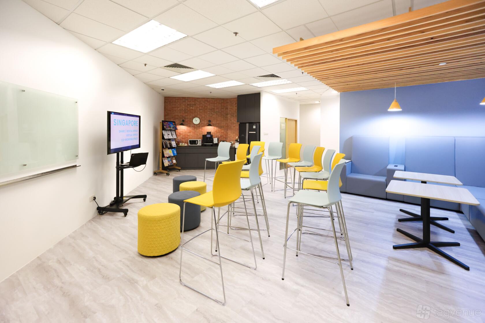 A coworking event space with bar-height chairs, ottomans, a whiteboard, and a wall-mounted screen at The Company Singapore.