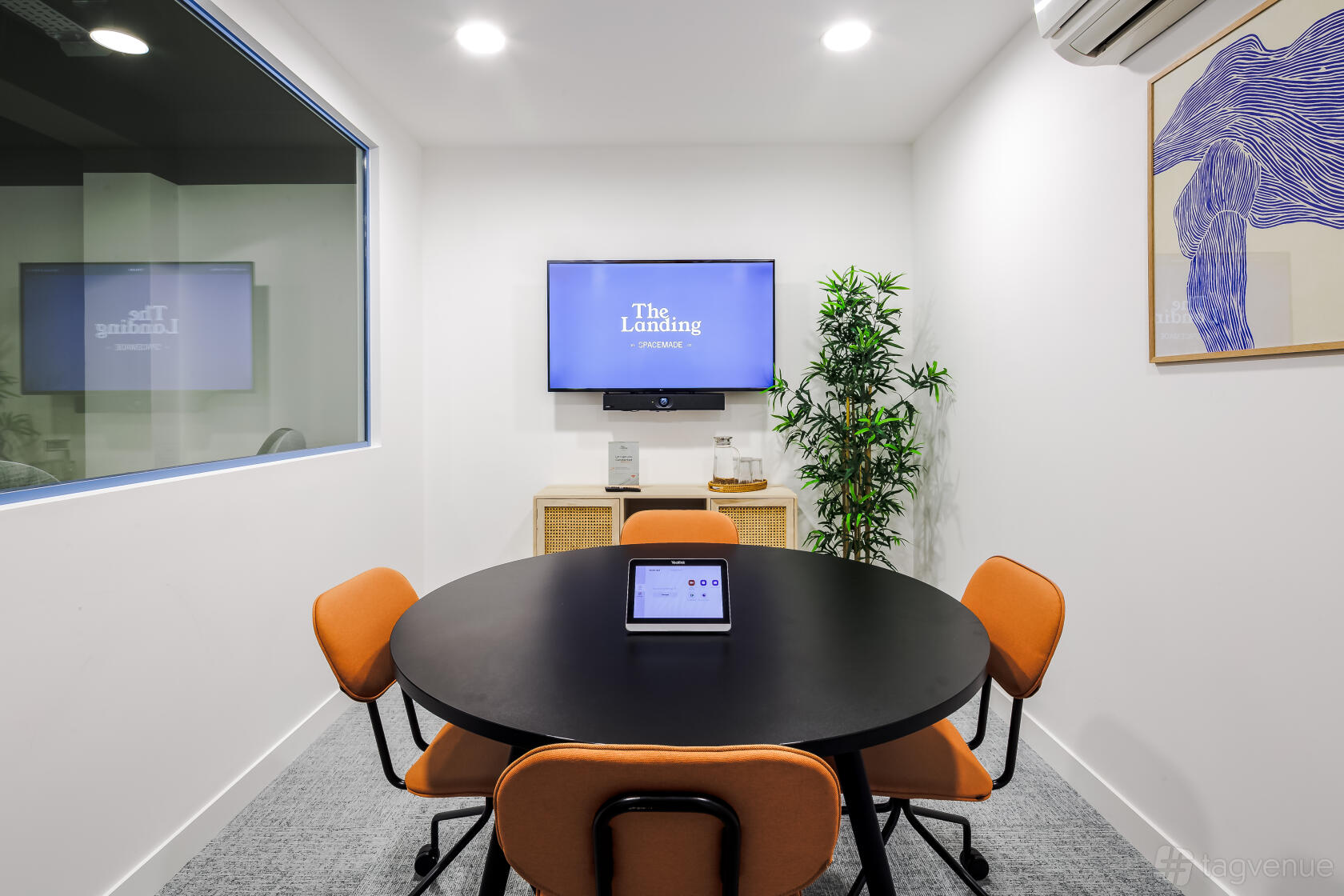 A meeting room with a round black table, orange chairs, wall-mounted screen, and potted plant at The Landing by Spacemade.