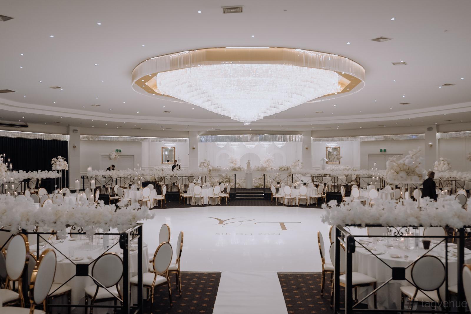 A ballroom with a grand central chandelier, white floral centerpieces, and round tables at Ultima Function Center.