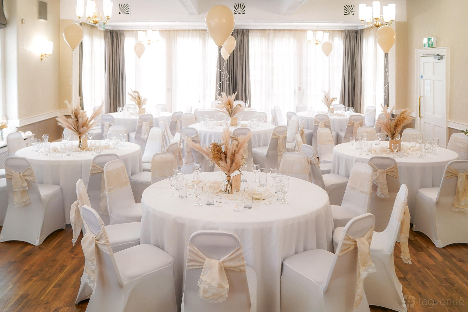 A ballroom with round tables covered in white linens, neutral chair sashes, and large windows at Flixton House.