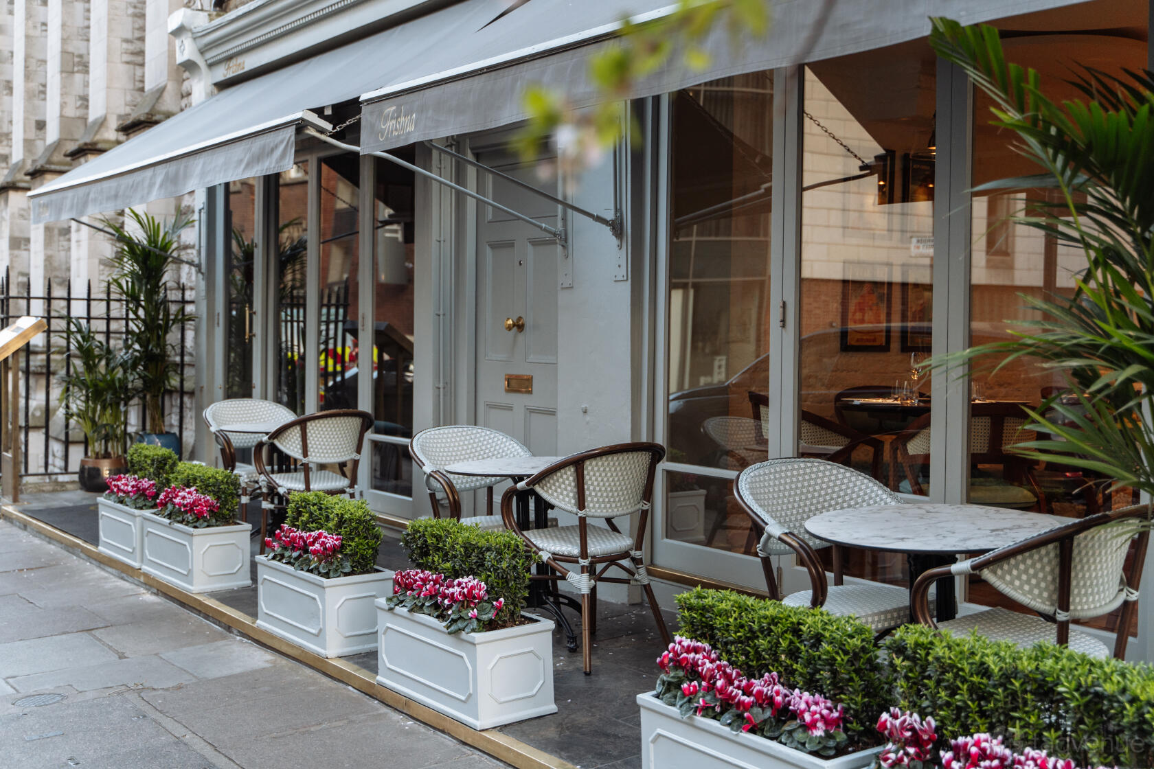 A restaurant with outdoor seating, rattan chairs, marble tables, and planters with flowers at Trishna.
