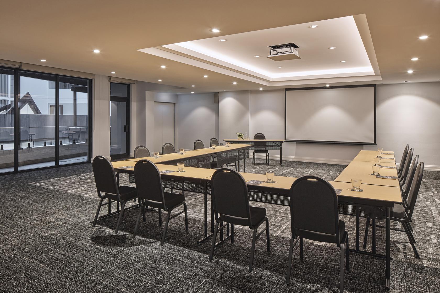 A meeting room with U-shaped tables, black chairs, a ceiling projector, and floor-to-ceiling windows at Rydges Sydney Central.