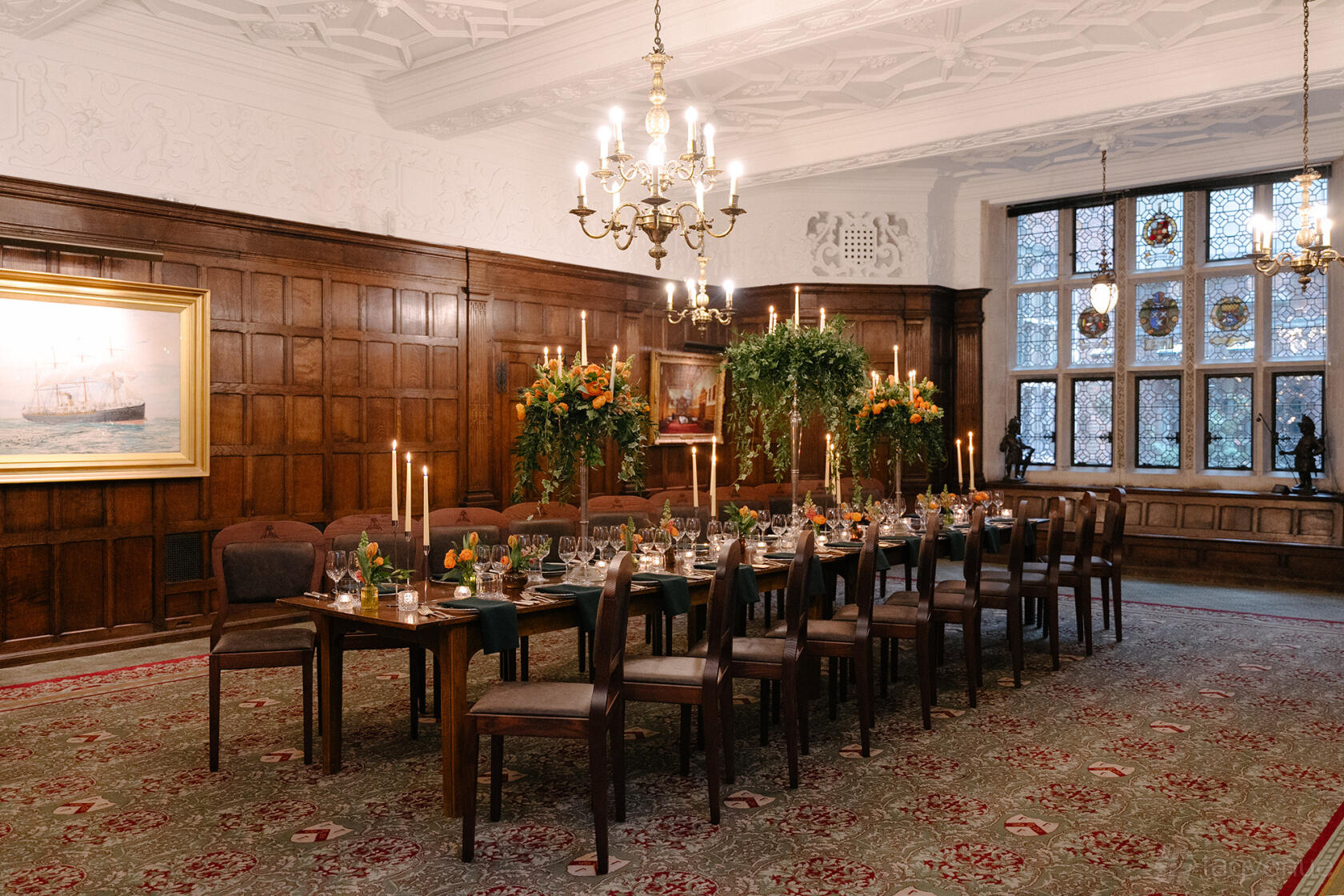 An event space in a hall with stained glass windows, wood-paneled walls, chandeliers, and banquet tables at Ironmongers' Hall.