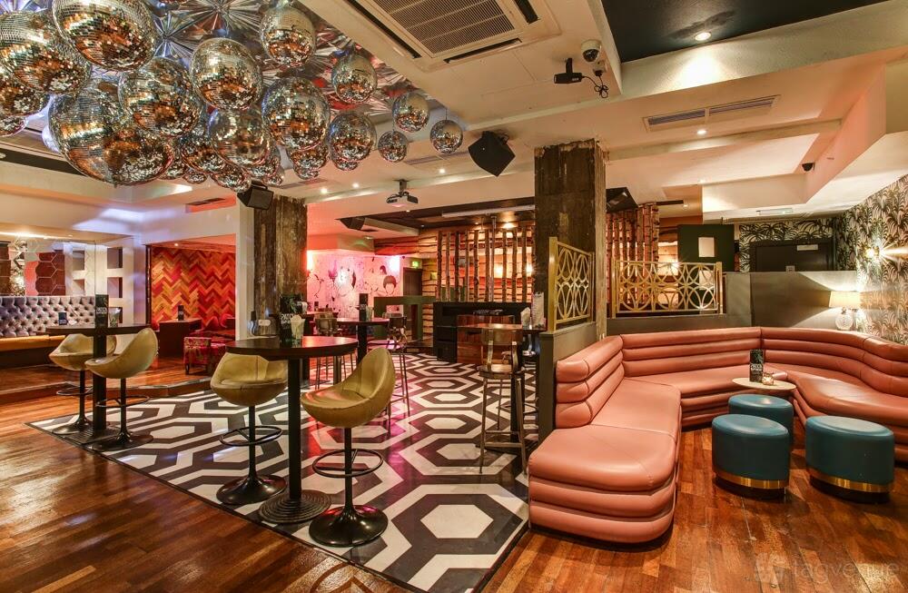 A bar with mirrored disco balls, geometric carpet, blush curved banquettes, and tall tables at Patch East.