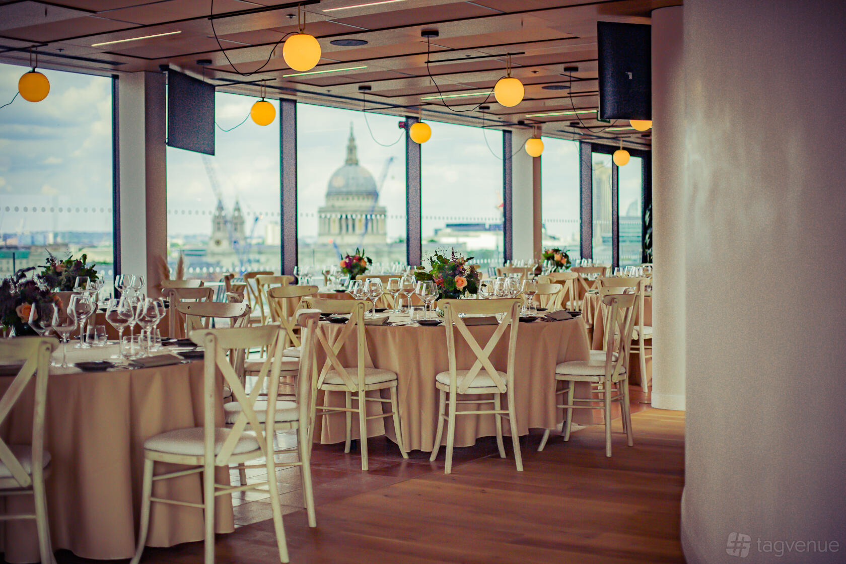 An event venue with round tables dressed in beige linens, white chairs, and floor-to-ceiling windows overlooking the city at Rose Court Events.