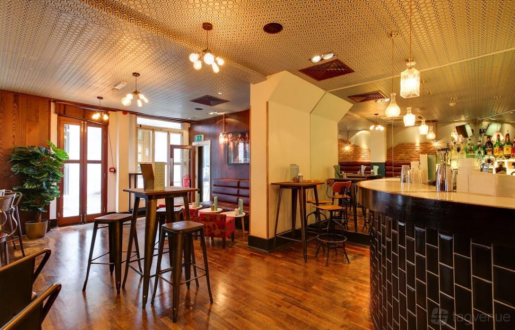 A function room with high tables, wooden flooring, pendant lighting, and a curved bar at Patch St Paul's