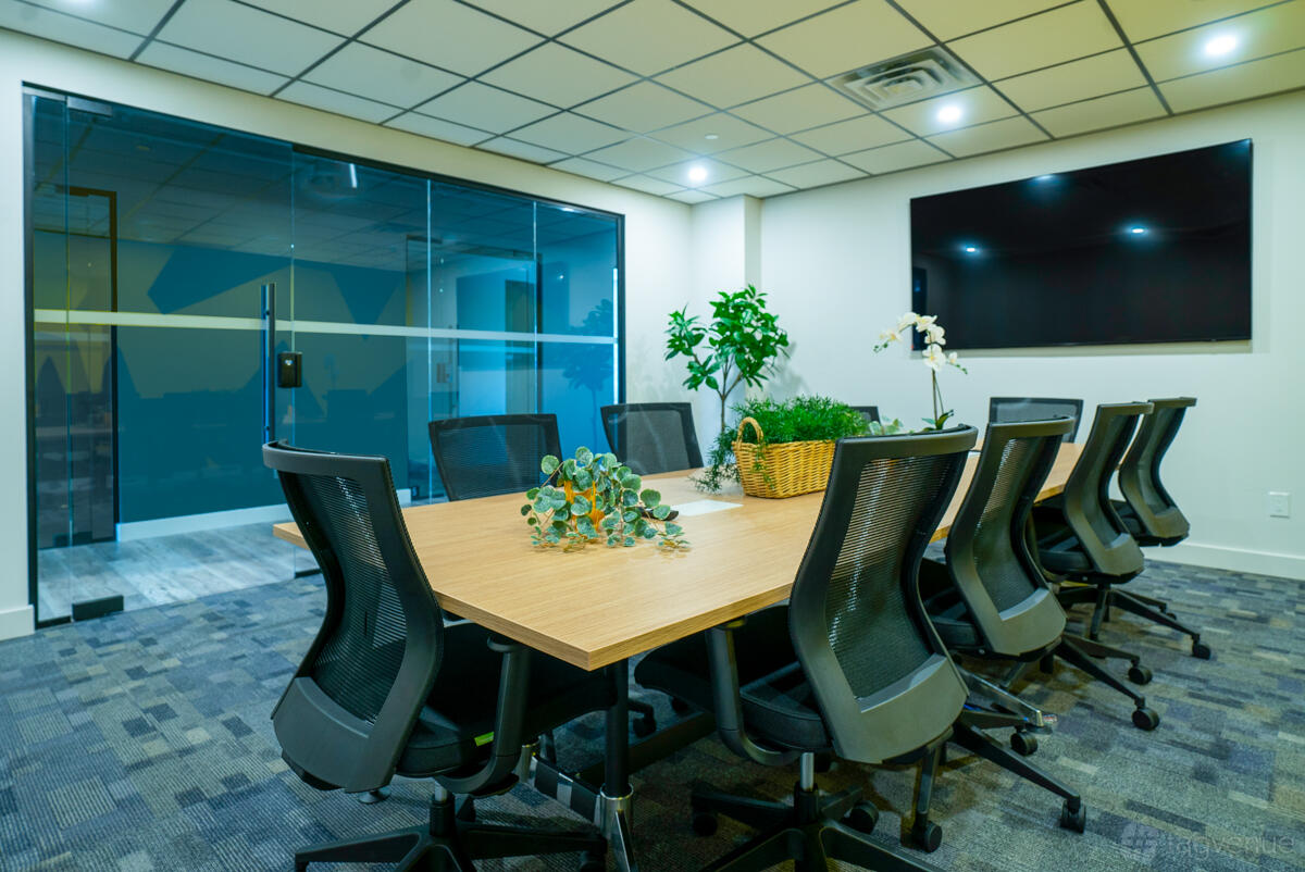 A meeting room with a long wooden table, black mesh chairs, large wall-mounted screen, and glass partition at Venture X Etobicoke.