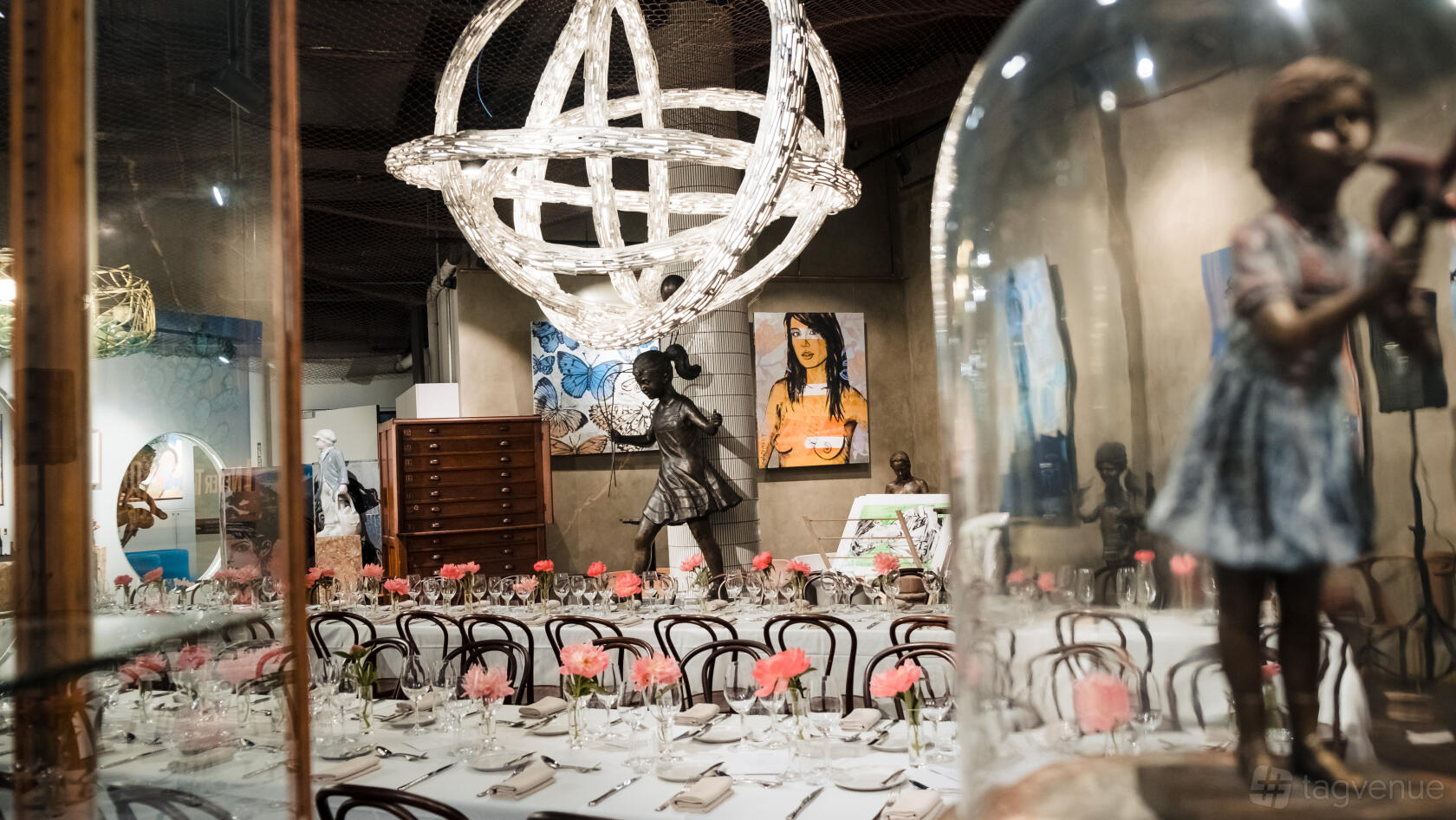 A gallery space with a long banquet table, pink floral centerpieces, artwork, sculptures, and a large orb chandelier at LK Hospitality.