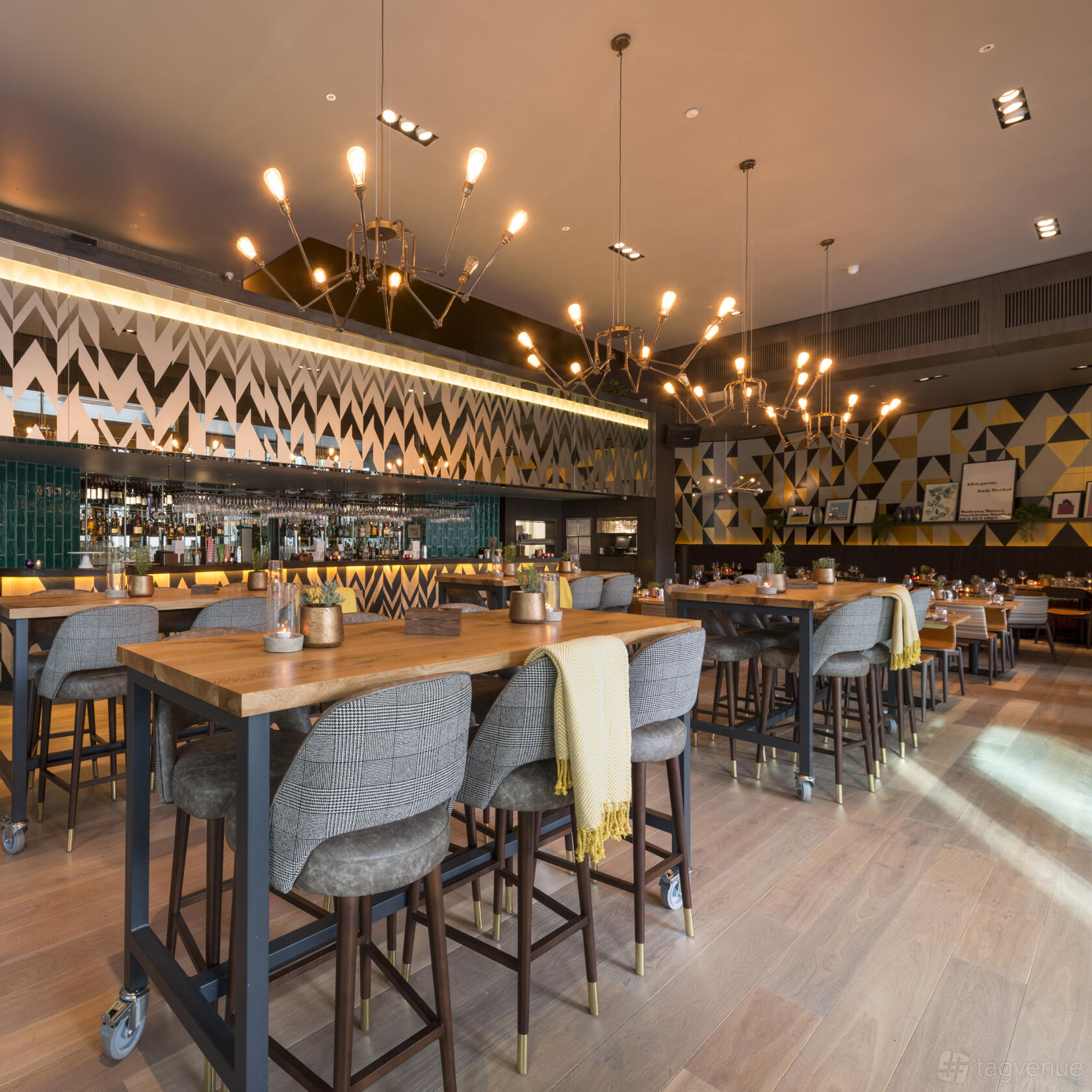 A bar in a restaurant with geometric wall patterns, velvet barstools, and wooden tables at The Refinery New Street Square.