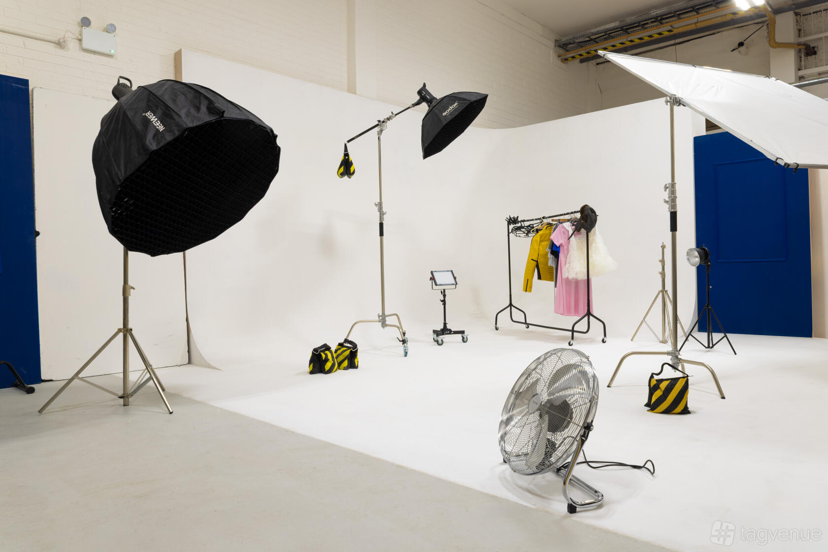 A photo studio with large softbox lights, white cyclorama walls, clothing rack, and a standing fan at Madsaint Studio.