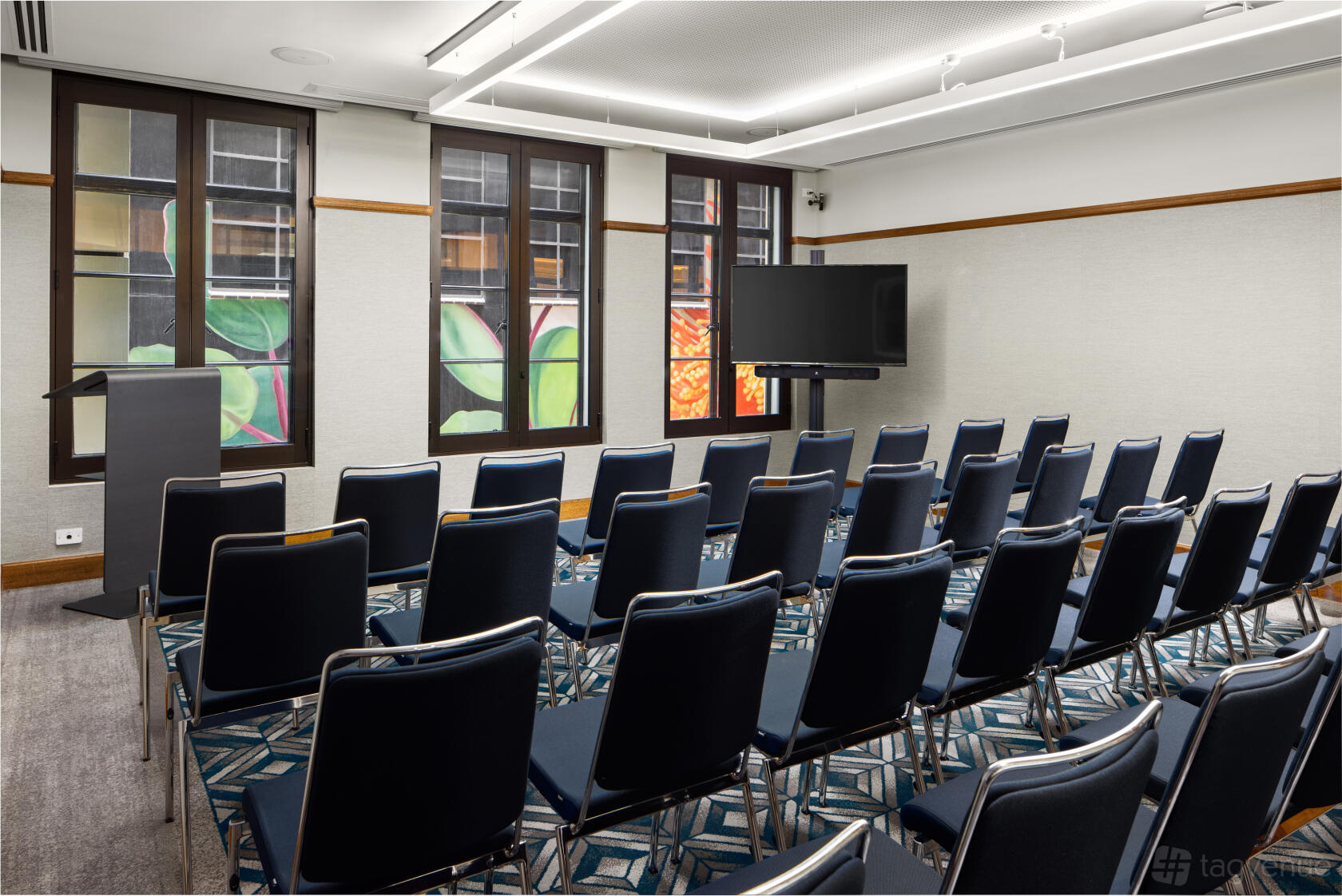 A meeting room with rows of navy chairs, patterned carpet, large windows, and a presentation screen at Hilton Melbourne Little Queen Street.