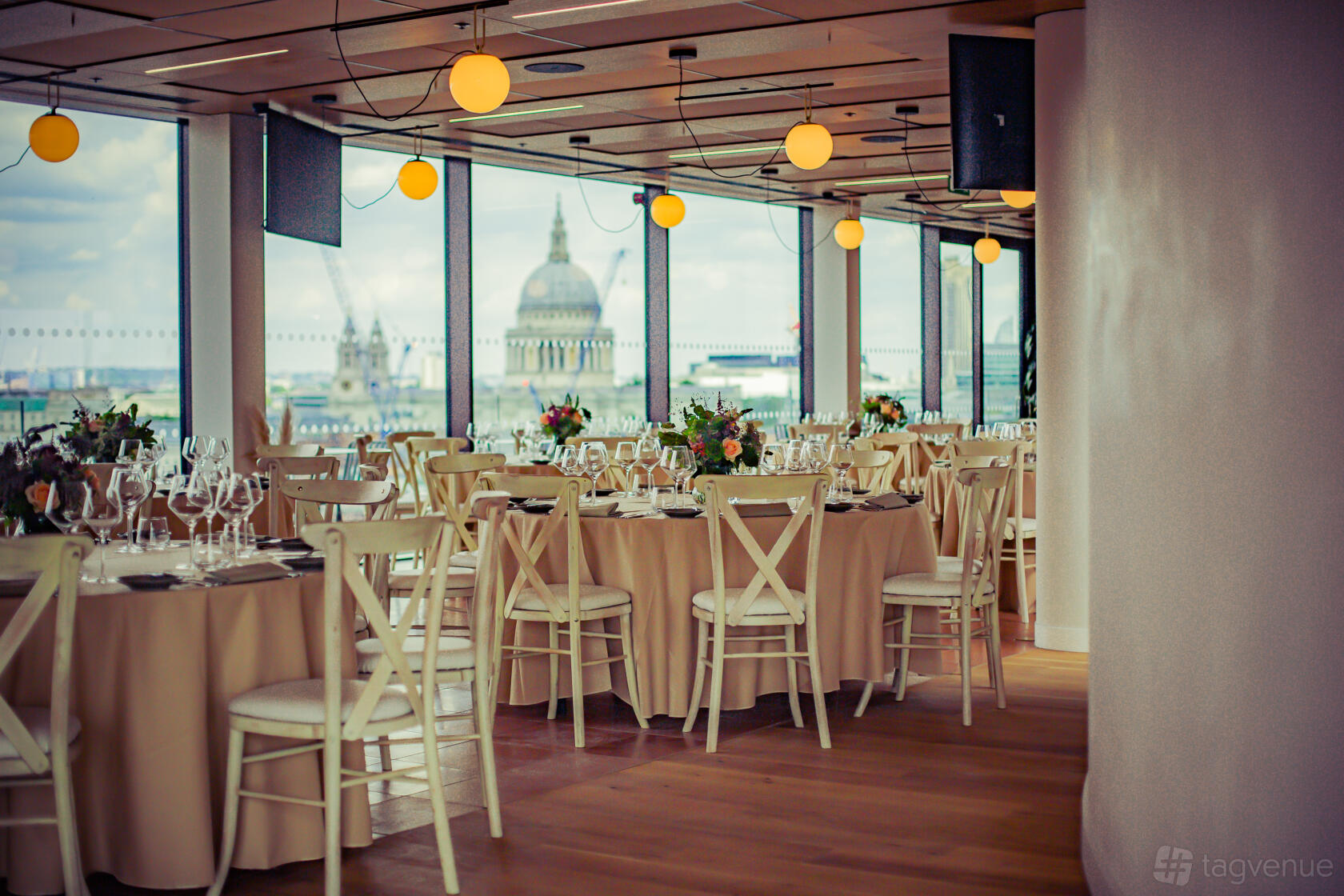 An event venue with floor-to-ceiling windows, round tables with beige linens, and city skyline views at Rose Court Events.