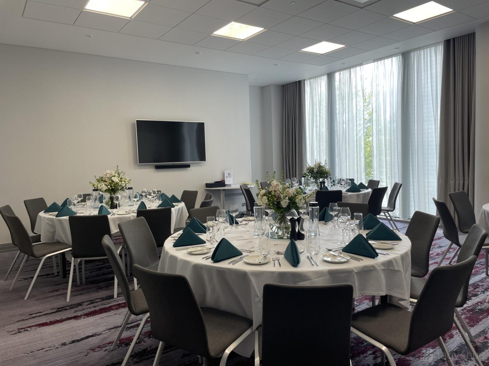 An event space with round tables set for dining, teal napkins, and floor-to-ceiling windows at Crowne Plaza London Docklands.