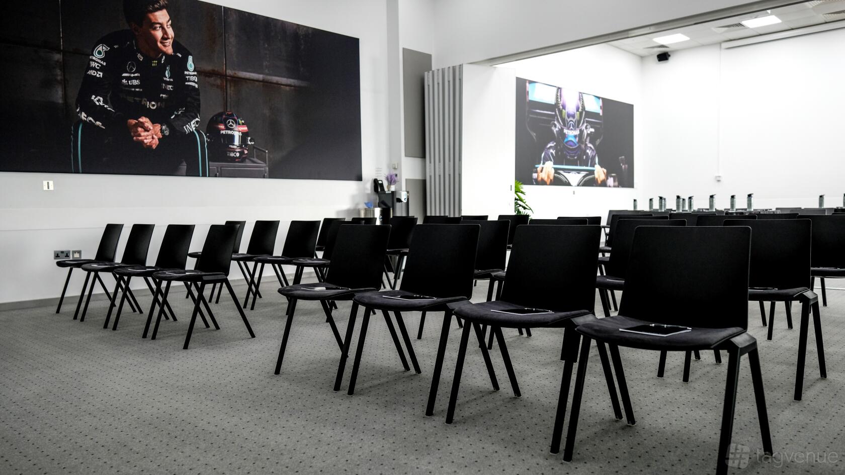 A meeting room with rows of black chairs, wall-mounted racing art, and a projector screen at Mercedes-Benz World.