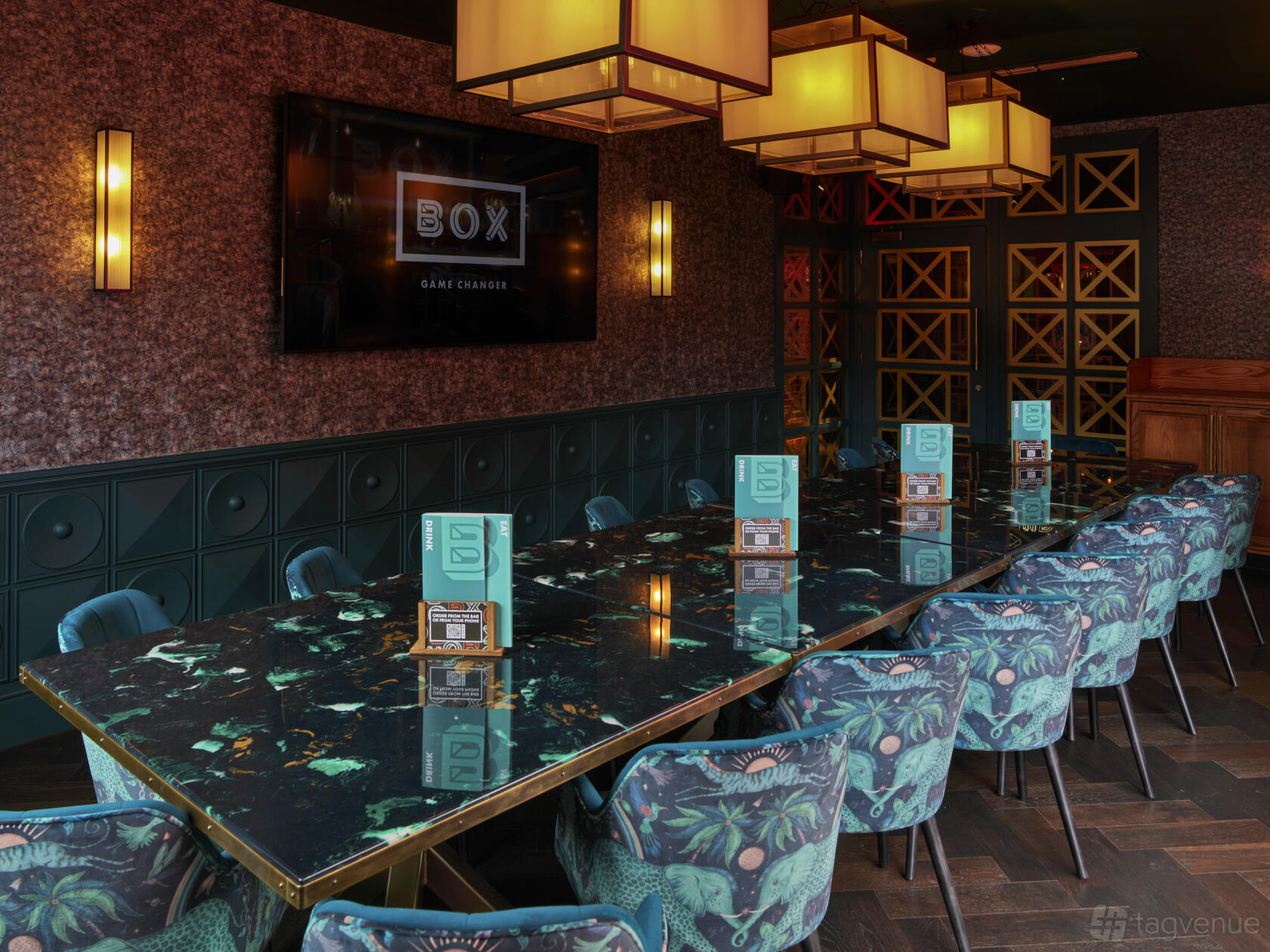 A dining room with a long marble table, velvet floral chairs, pendant lighting, and a large screen at Box Brindley Place.