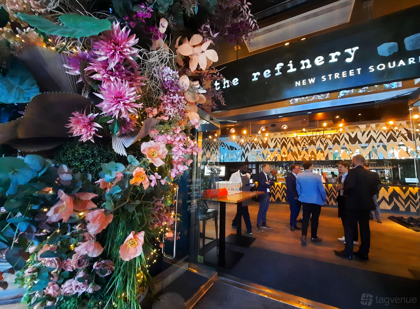 A bar with a geometric patterned counter, pendant lighting, and a floral entrance at The Refinery New Street Square.