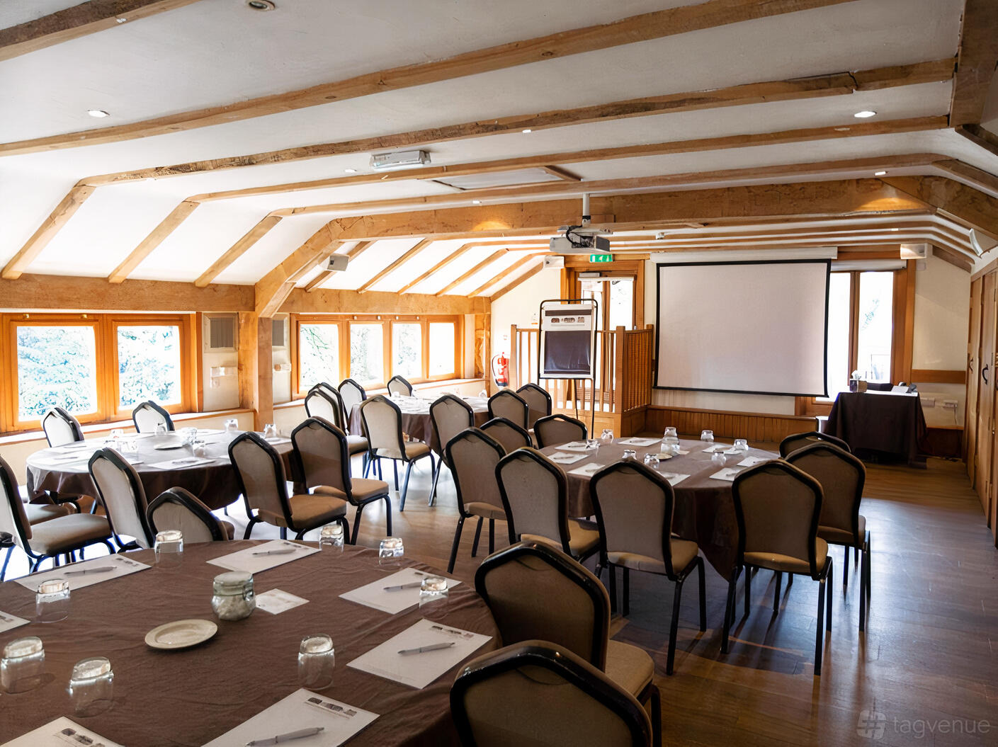 A function room with large windows, exposed wooden beams, round tables, and a projector screen at Tewinbury Farm Hotel.