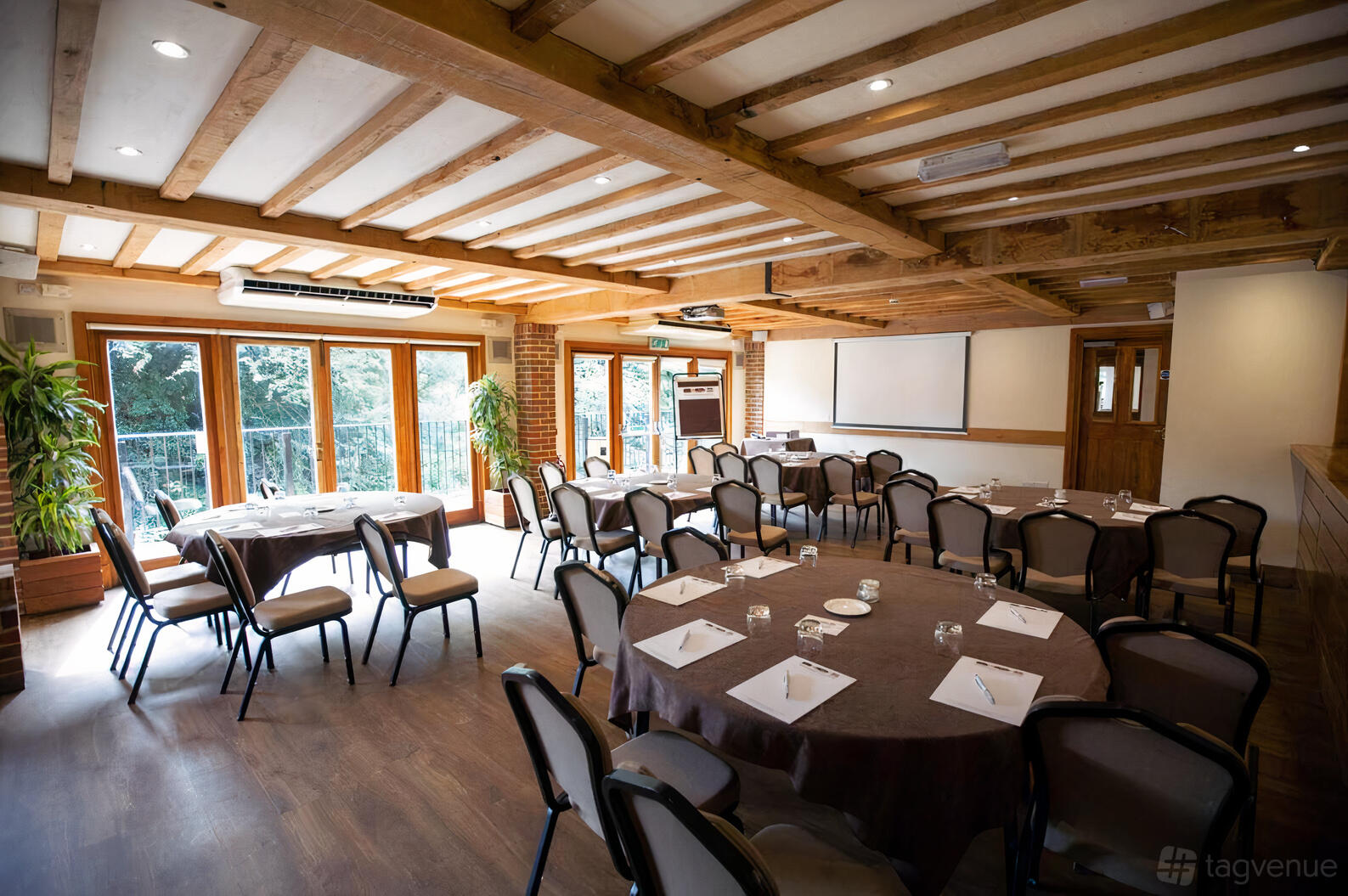 A function room with exposed wooden beams, large windows, and round tables set for a meeting at Tewinbury Farm Hotel.