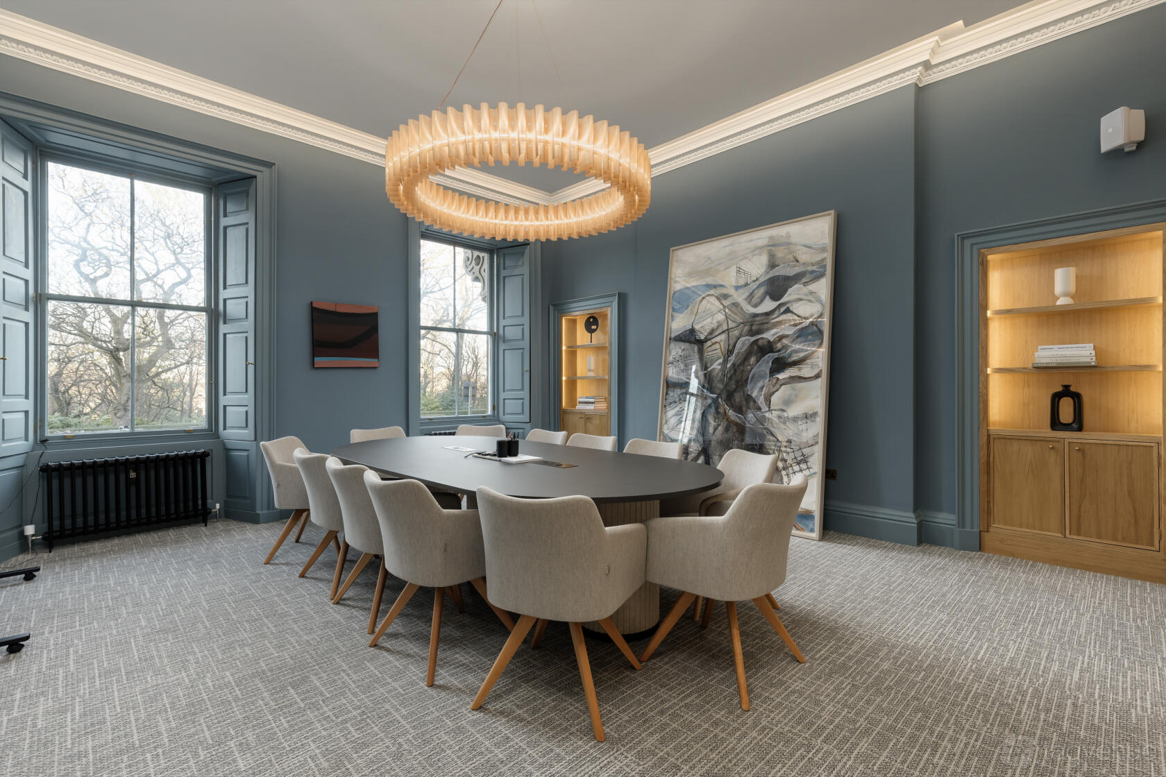 A boardroom with a long oval table, cushioned chairs, large windows, and a circular chandelier at The Auction Rooms.